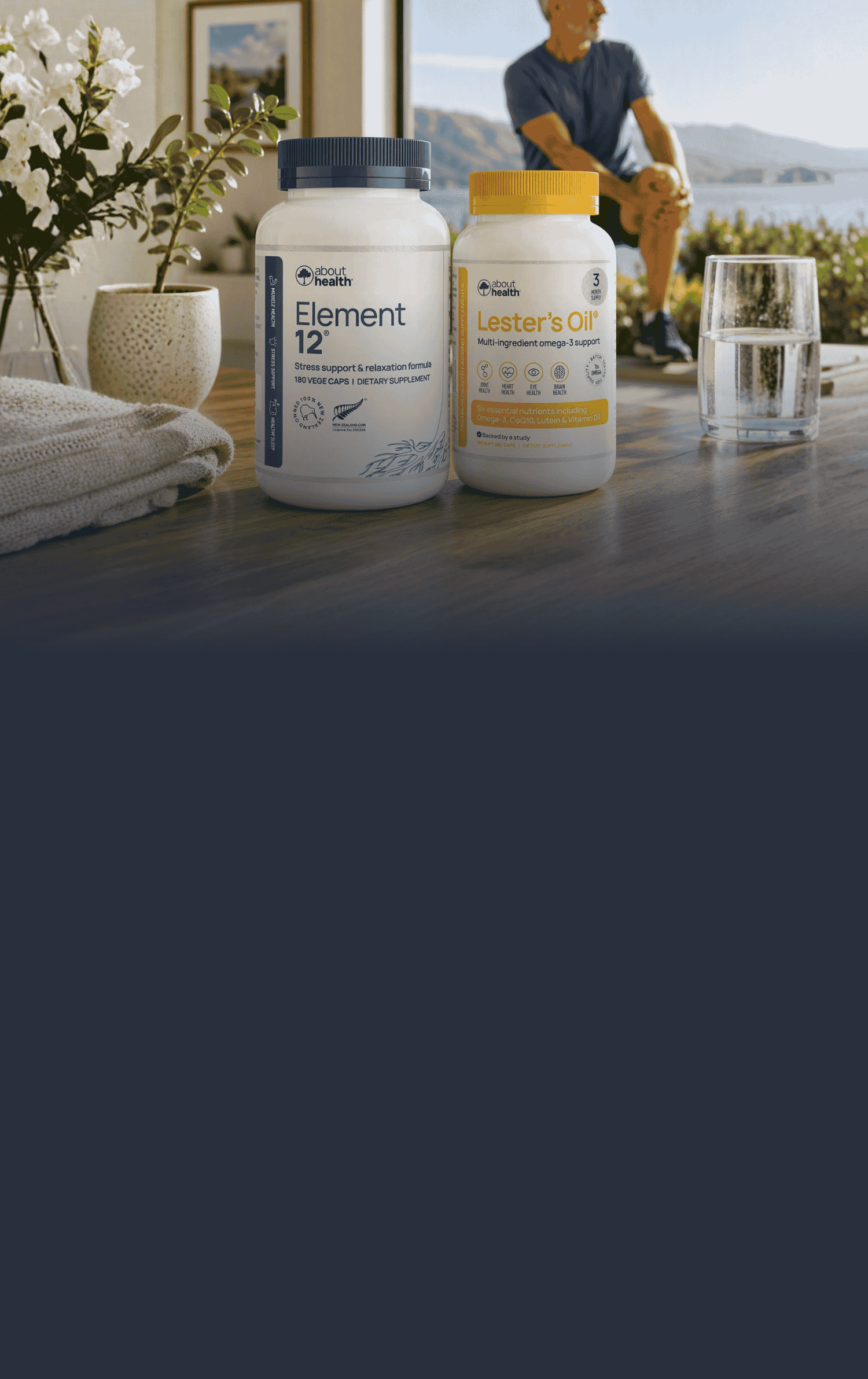 Two supplement bottles from Vitality Living are displayed on a dark wooden table in a lifestyle setting with a man sitting in the background near a window overlooking water.