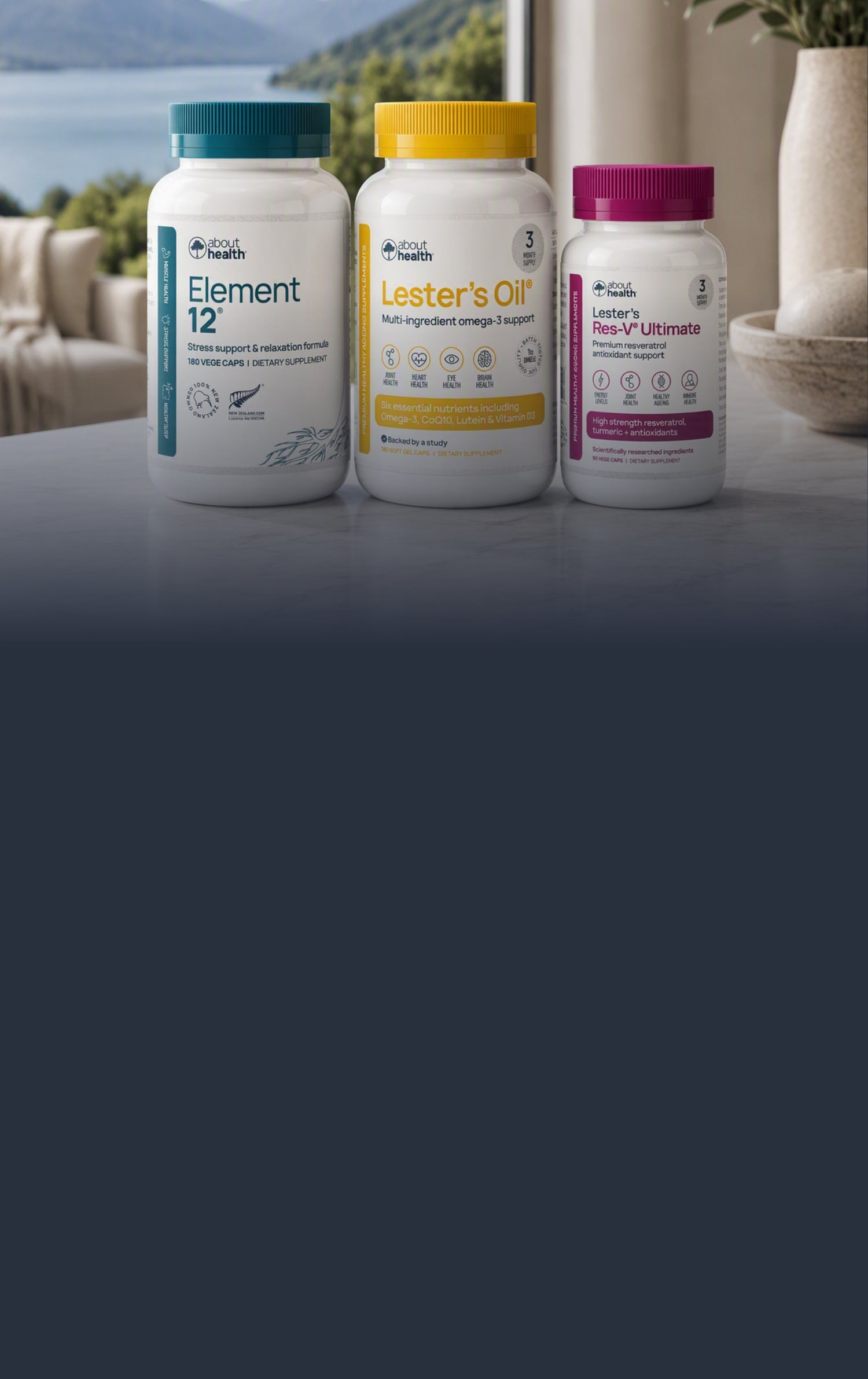 Three supplement bottles from Good Health are lined up on a dark tabletop in front of a blurred indoor background with a window view. The bottles are white with colored caps and labels: teal for Element 12, yellow for Lester's Oil, and purple for Lester's Res-V Ultimate