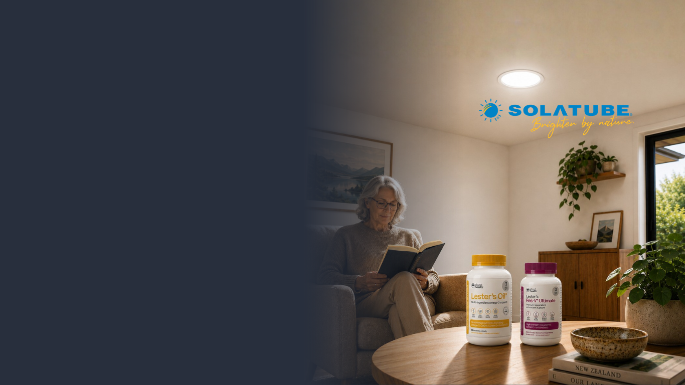A lifestyle scene in a cozy living room showing an elderly woman reading a book in an armchair, with SOLATUBE  skylight glowing from a recessed ceiling light above. In the foreground, Lester's Oil and Res-V Ultimate are placed on a wooden table next to a window.