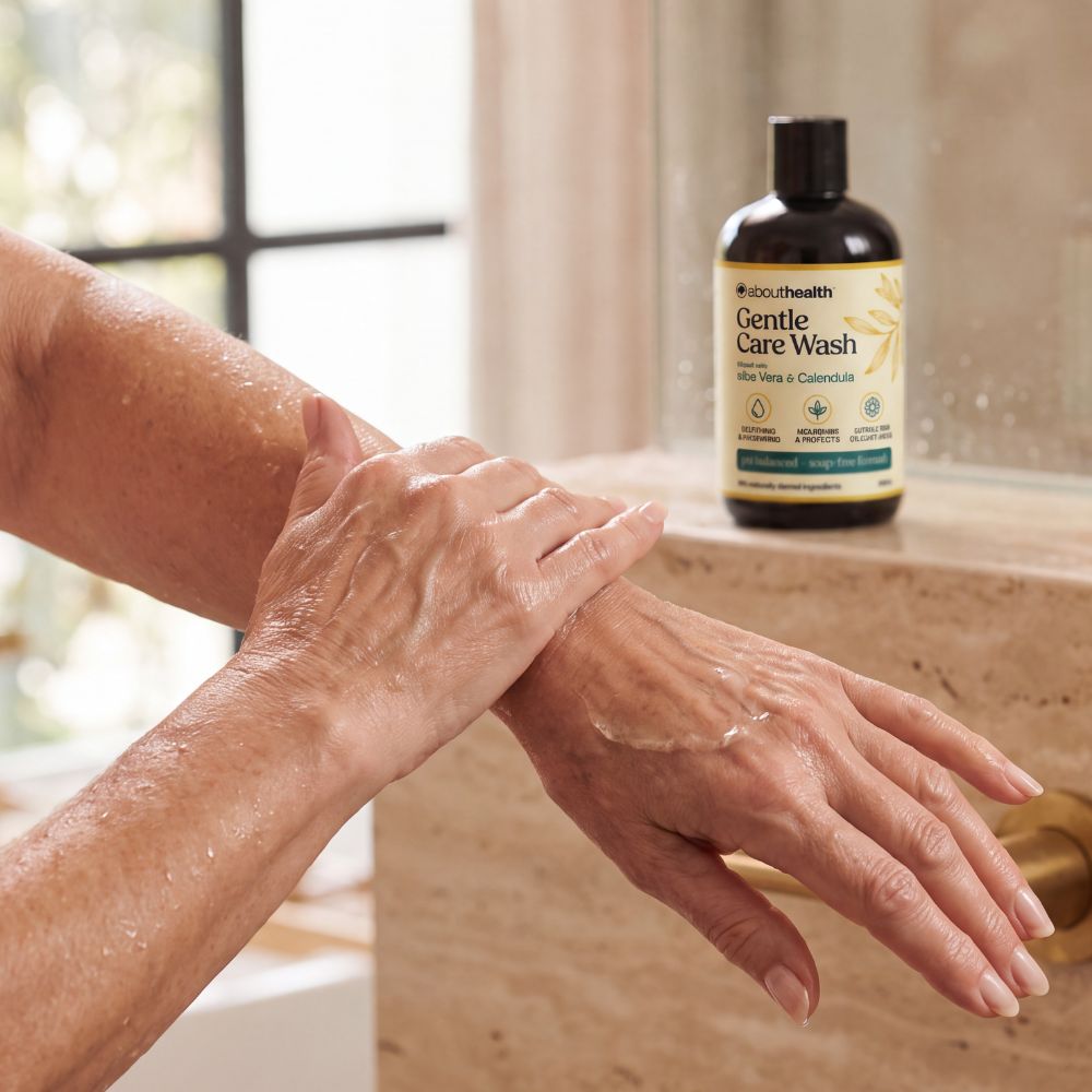 A person's hands lathered with soap wash are shown in a bathroom setting next to a bottle of About Health Gentle Care Wash on a beige stone counter.