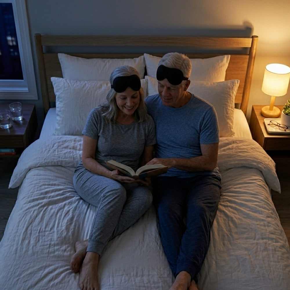 Couple sitting on a bed reading a book together, wearing eye masks.