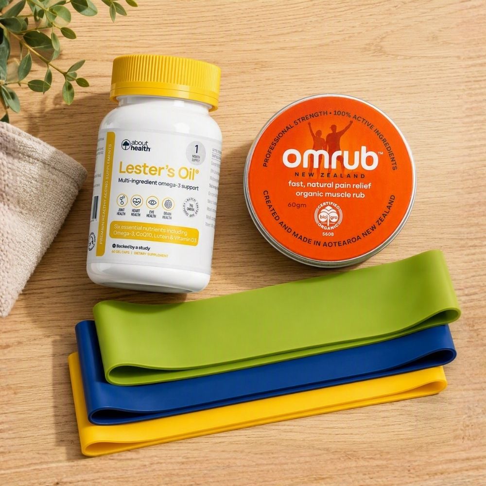 A wellness product set arranged on a light wood surface, including a white bottle of about:meals Lester's Oil with a yellow cap, an orange tin of omrub Muscle Relief, and three resistance bands in green, blue, and yellow.