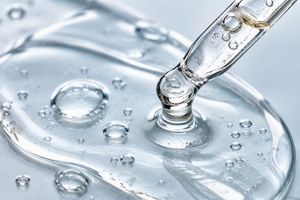 A close-up of a clear glass dropper tip dispensing glycerin onto a glossy surface with visible water droplets and reflections.