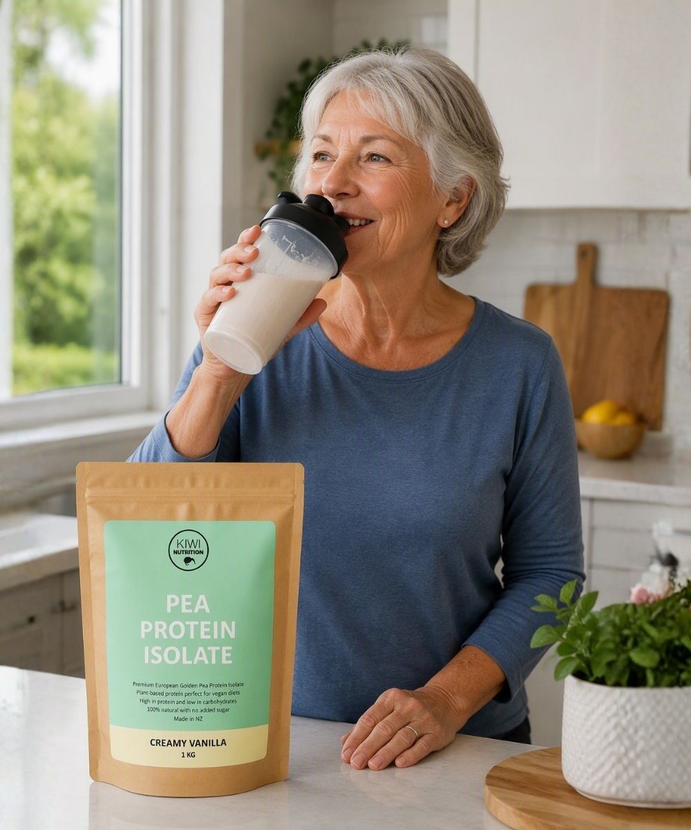 An older woman in a blue shirt stands in a bright kitchen, drinking from a white shaker bottle. In the foreground on the counter is a stand-up pouch of pea protein isolate labeled 'PEA PROTEIN ISOLATE' and 'CREMIY VANILLA'.