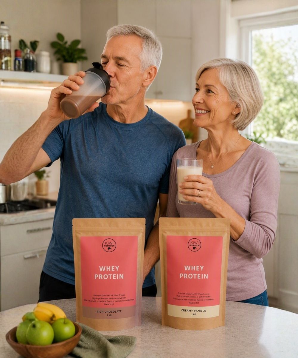 An older couple in a bright kitchen, with the man drinking from a shaker bottle and the woman holding a glass of protein shake. Two stand-up pouches of Whey Protein are displayed on the counter in front of them, one labeled 'Rice Chocolate' and the other 'Creamy Vanilla'.
