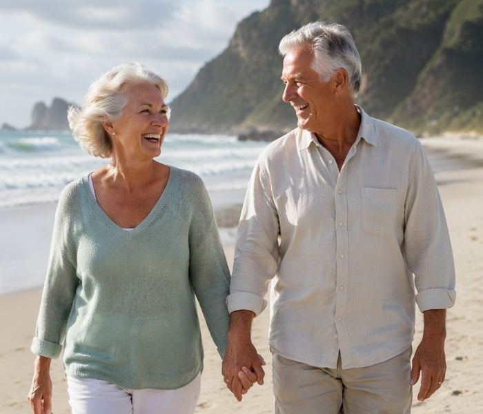 An older couple holding hands and smiling as they walk along a sandy beach with ocean waves and rocky cliffs in the background in New Zealand