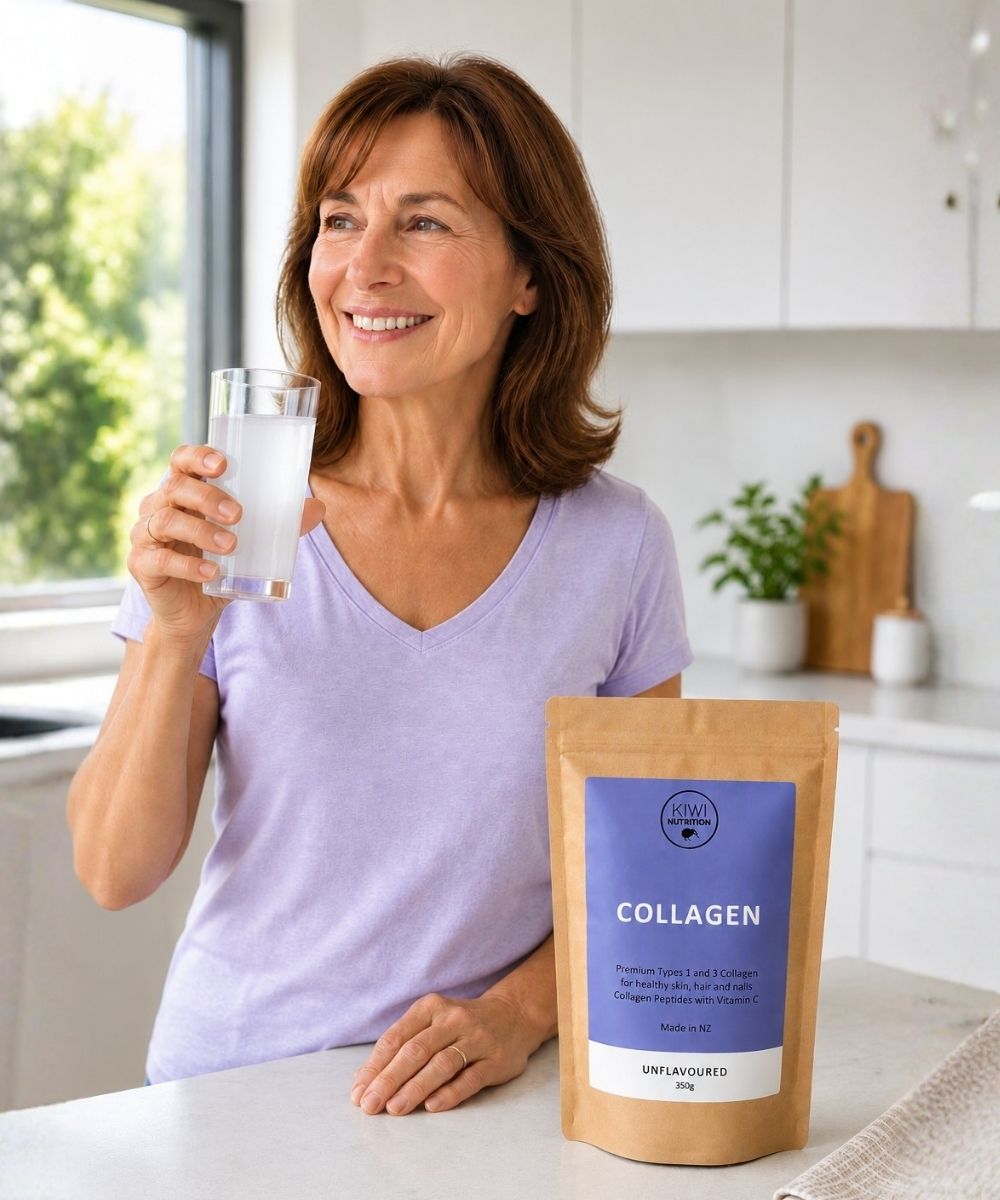 A smiling woman in a light purple V-neck shirt stands in a bright kitchen holding a glass of milk, with a stand-up pouch of collagen powder placed on the counter in front of her. The pouch has a kraft brown exterior and a blue label reading 'COLLAGEN'.