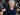 A smiling older woman with gray hair is exercising on a gym machine, wearing a navy blue short-sleeve athletic shirt performing pec flys on a machine.