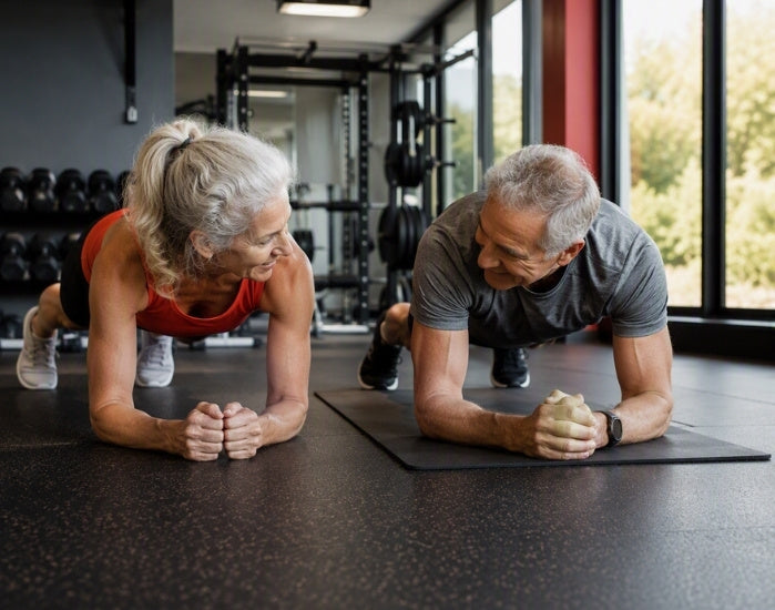 An older couple doing a plank exercise in a modern gym. The woman on the left wears a red sports bra and black shorts, while the man on the right wears a gray t-shirt and black athletic pants.