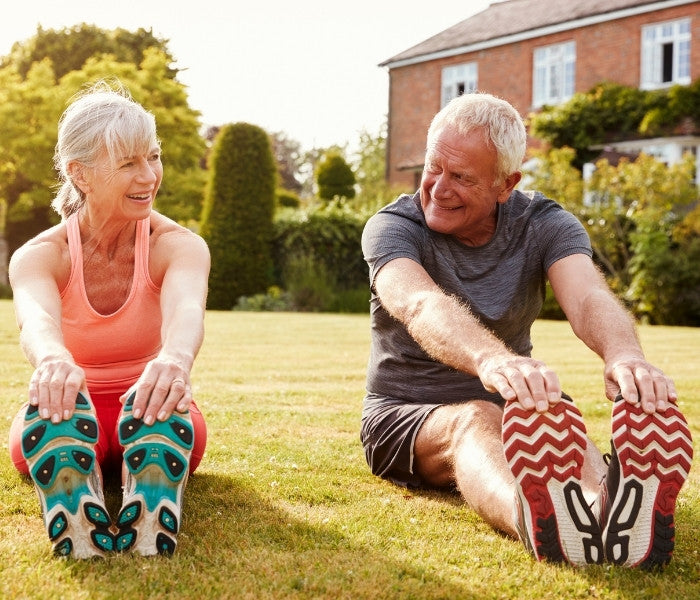 Active senior couple stretching outdoors on grass with a house in the background.