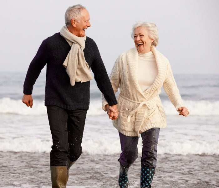 Two elderly people walking on a beach holding hands, smiling.